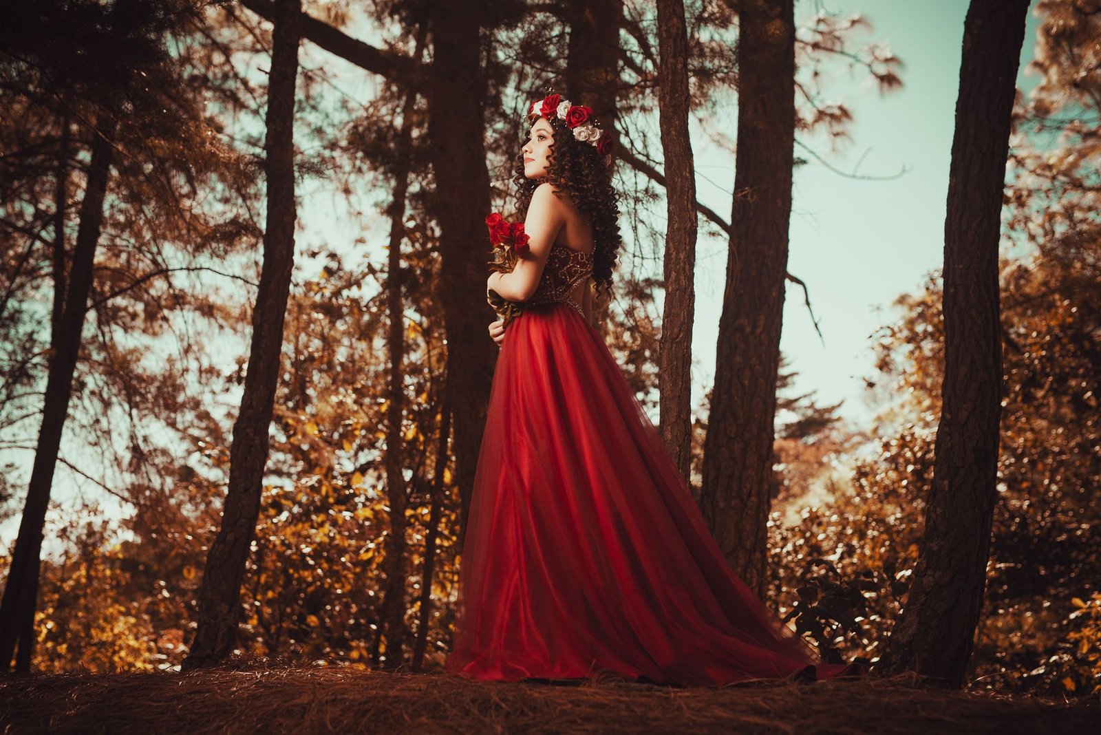 Woman in a flowing red dress and floral crown standing in a forest, evoking Persephone from Greek myth