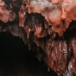 Red and brown stalactites hang from the ceiling of a dark cave, evoking Aeneas’s descent into the underworld
