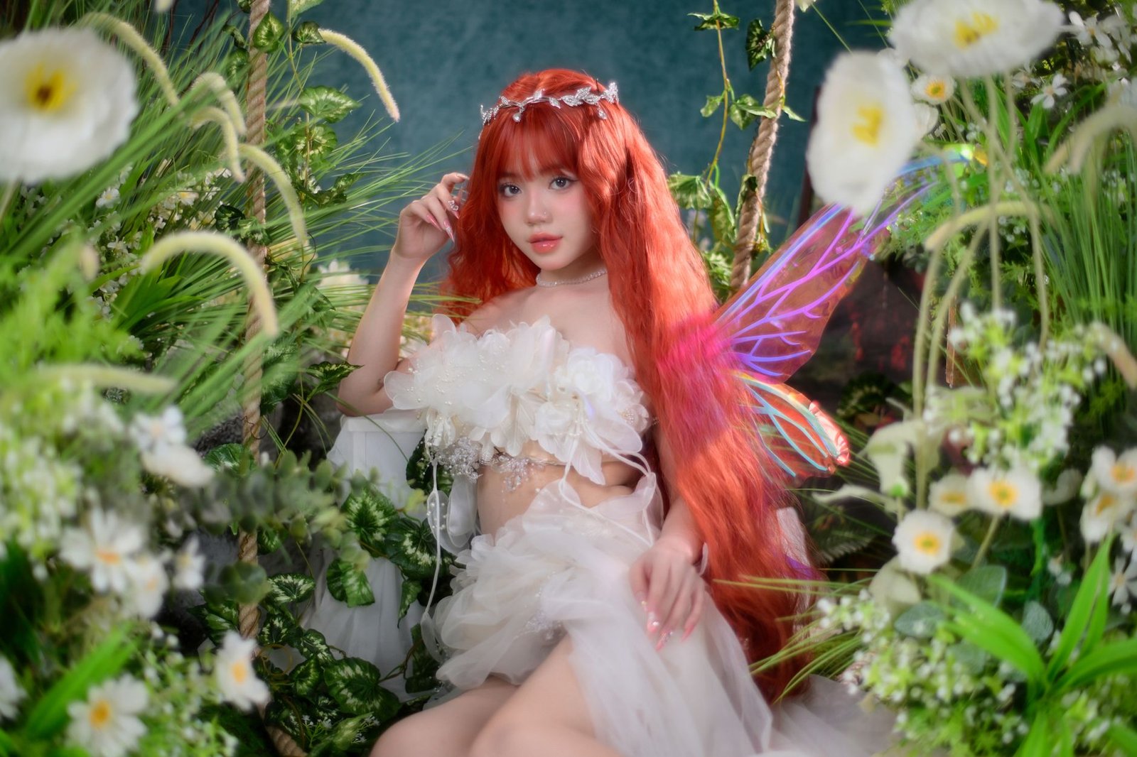 Red-haired fairy with wings standing among flowers and greenery in a cosplay scene