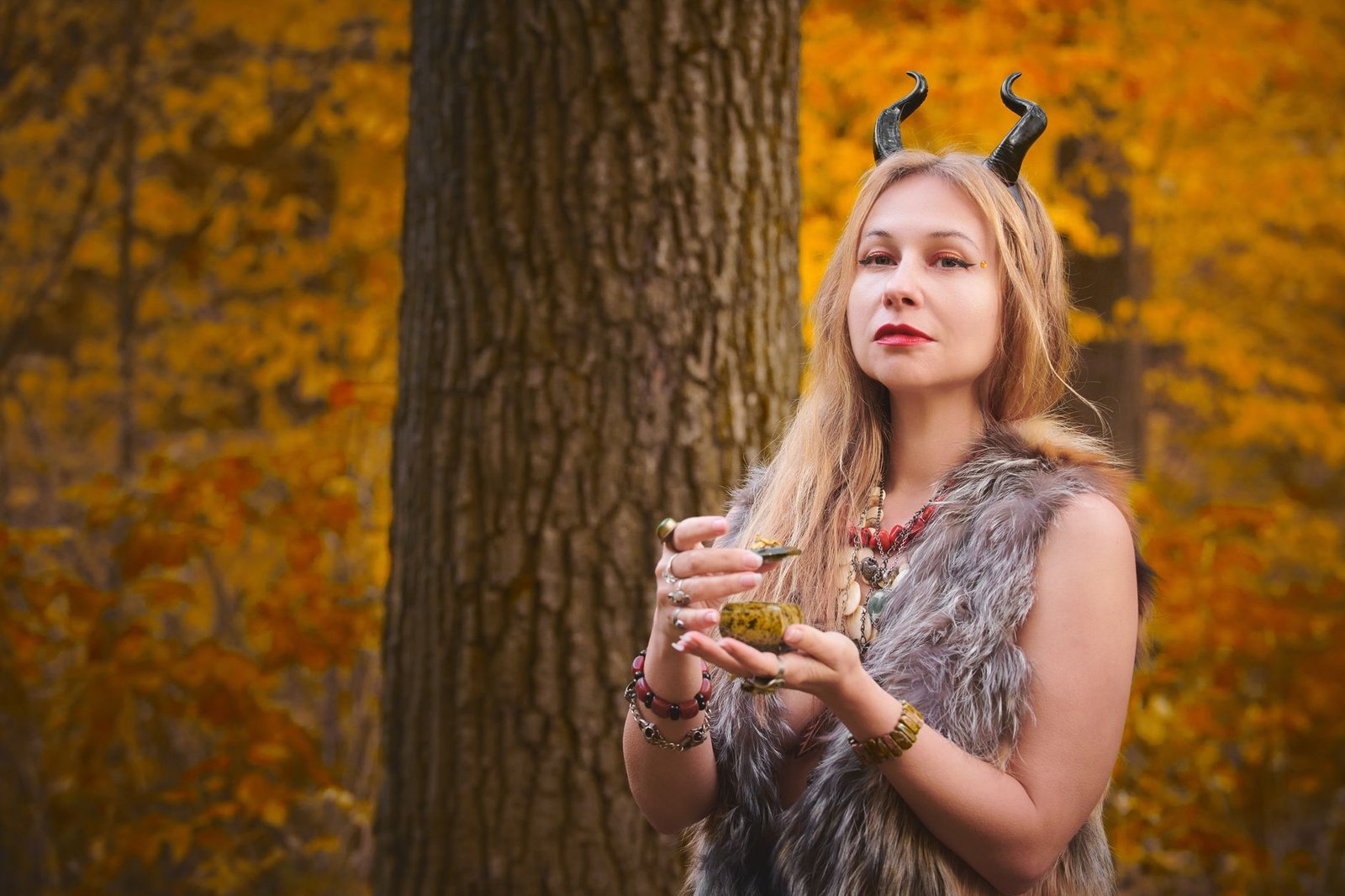 Young woman in a fur vest and horn headband holding a potion bottle in an autumn forest, evoking Arianrhod mythology