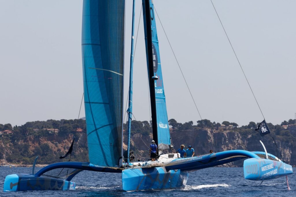 Blue trimaran sailboat racing on open water near a rocky coastline, evoking Atalanta's race and the golden apples