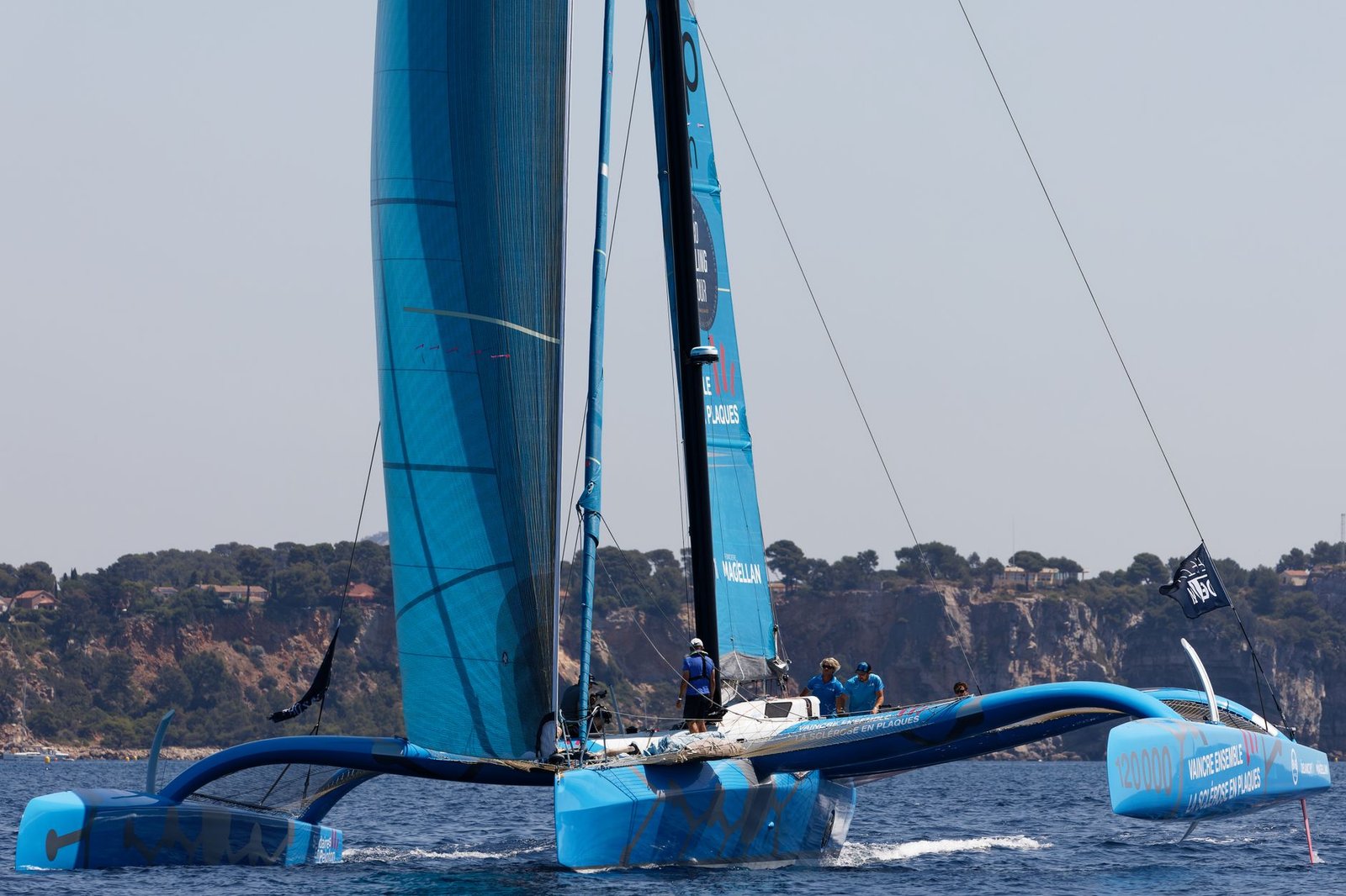 Blue trimaran sailboat racing on the ocean near a rocky coastline, evoking Atalanta's race and golden apples