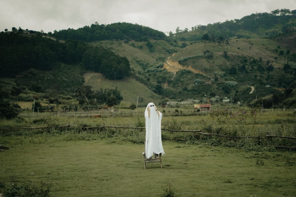 Person dressed as a ghost standing on a stool in a grassy field with mountains in the background, evoking a banshee