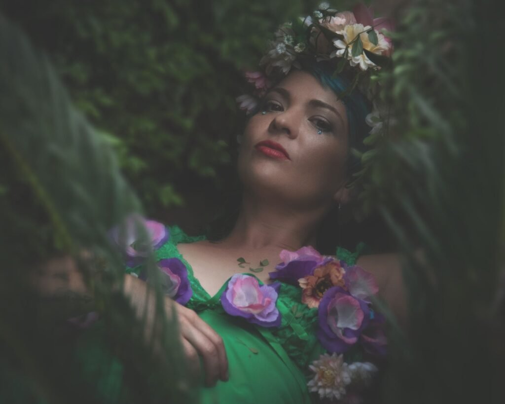 Portrait of a woman in a floral crown and green dress surrounded by lush greenery, evoking Blodeuwedd from Welsh myth