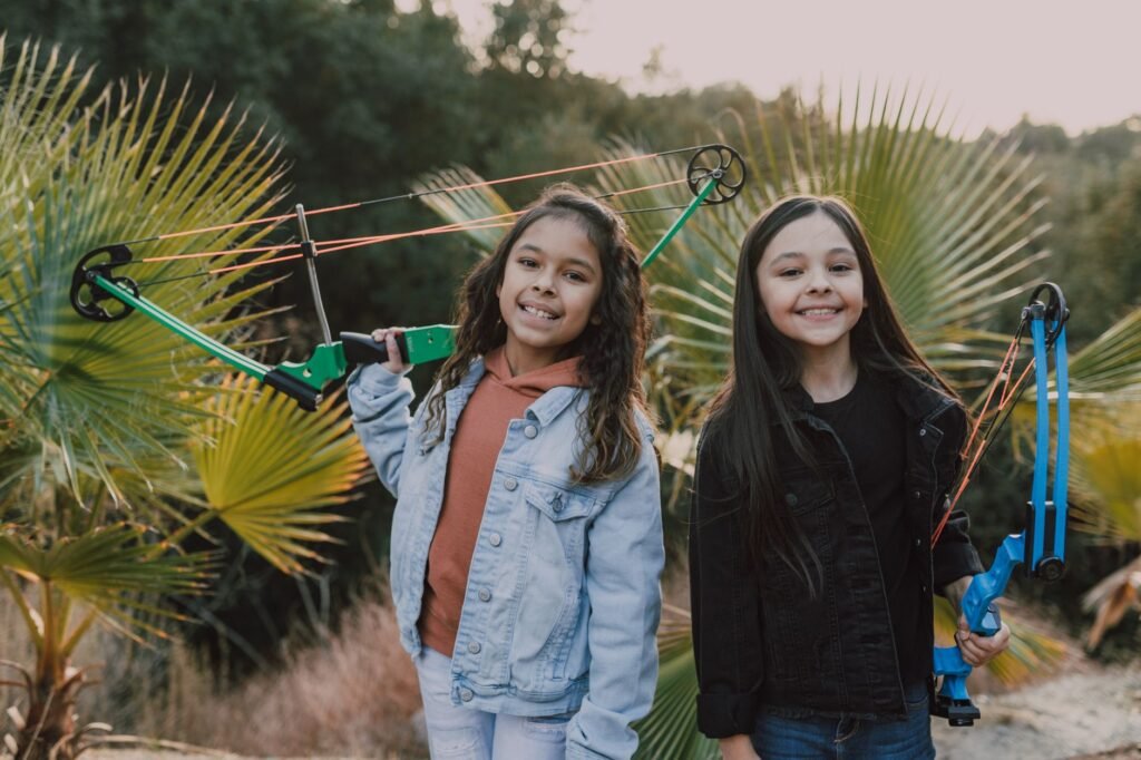 Two smiling girls holding archery bows outdoors in a natural setting