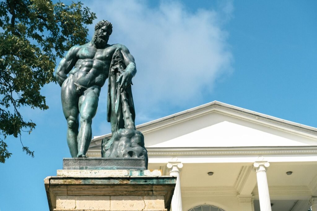 Bronze statue of a muscular man on a pedestal against a blue sky and classical building, evoking Cronus from Greek myth
