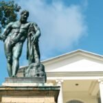 Bronze statue of a muscular man standing on a pedestal before a classical building with columns, representing Cronus
