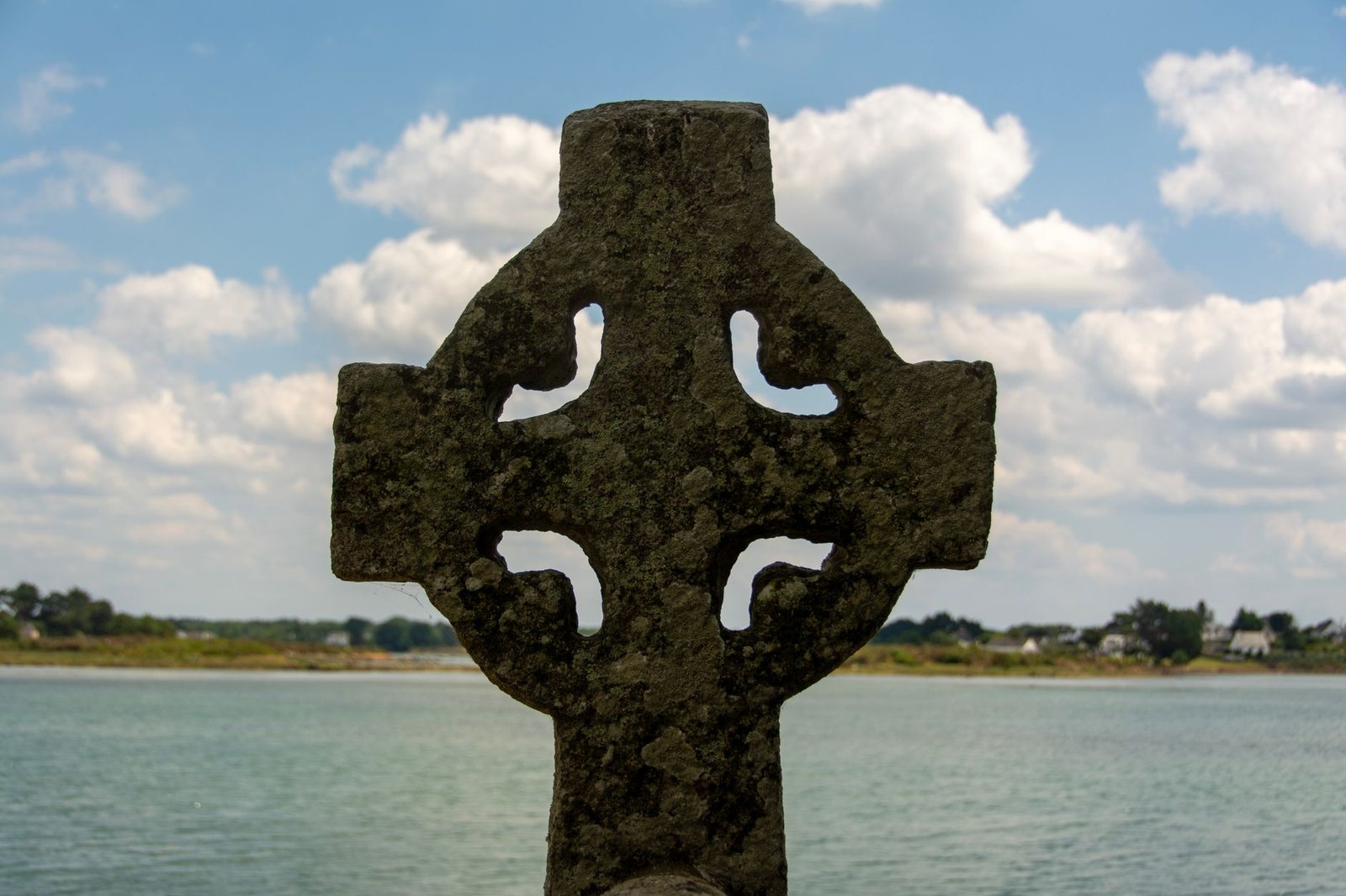Ancient Celtic cross monument overlooking a calm lake beneath a partly cloudy sky