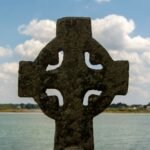 Ancient Celtic cross monument with knotwork overlooking a calm lake under a partly cloudy sky