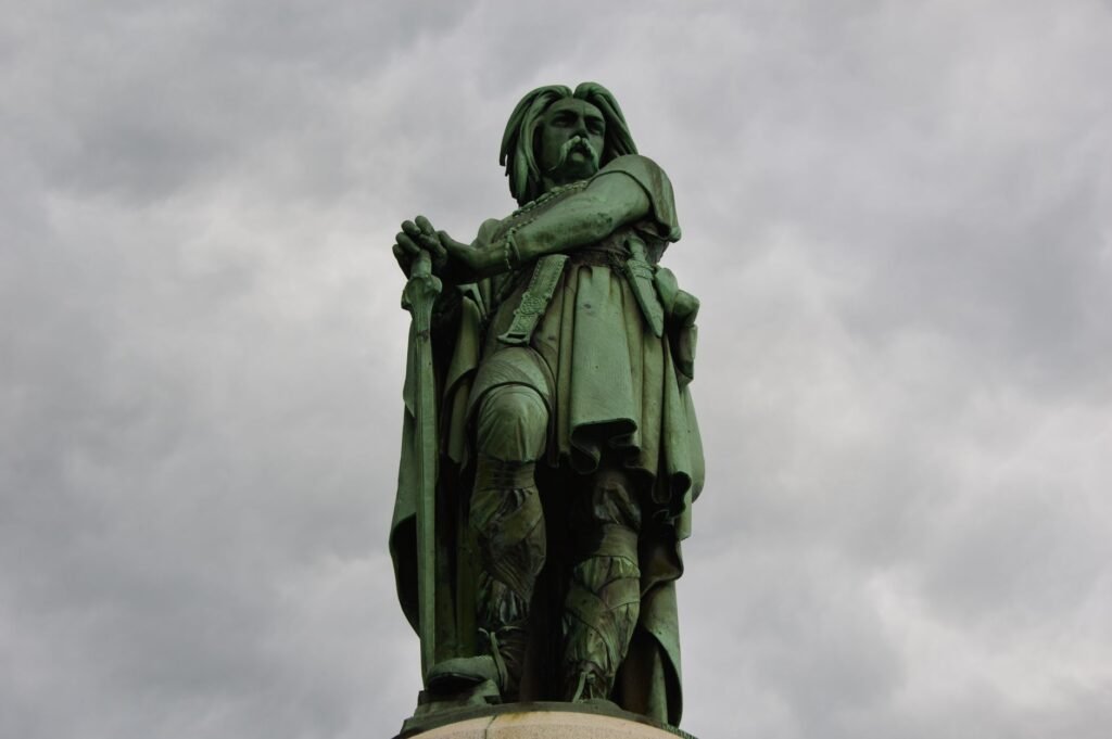 Statue of a historical figure holding a sword against a cloudy sky, representing the chief god in Celtic mythology