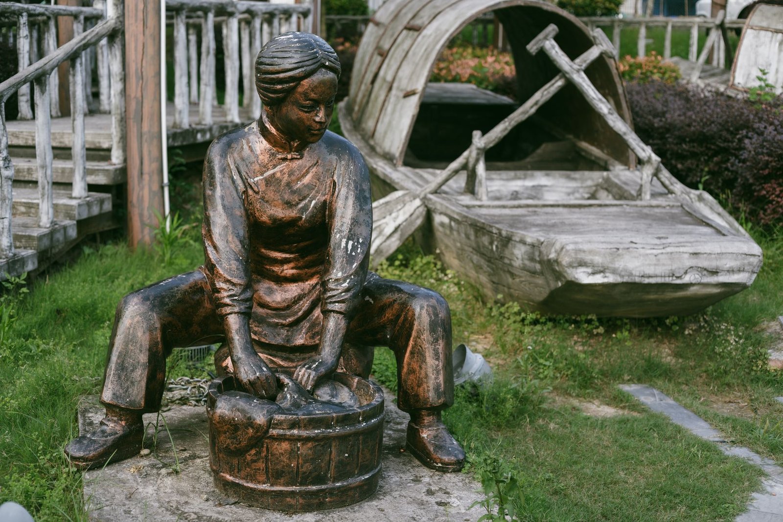 Bronze statue of a woman washing clothes outdoors beside a wooden boat and rustic fence