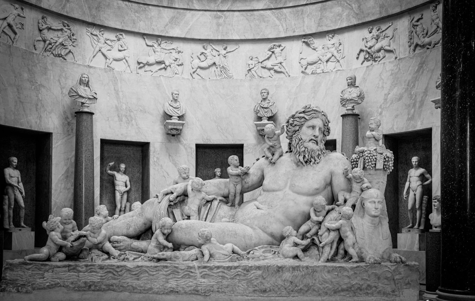 Black and white photo of a reclining Roman god statue surrounded by smaller figures in a museum display