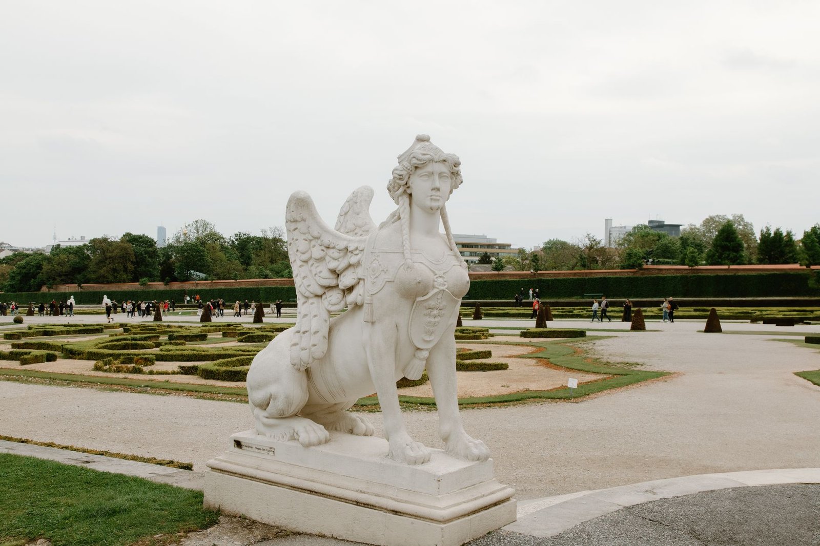 White stone sphinx statue in a formal garden with trimmed hedges and pathways.