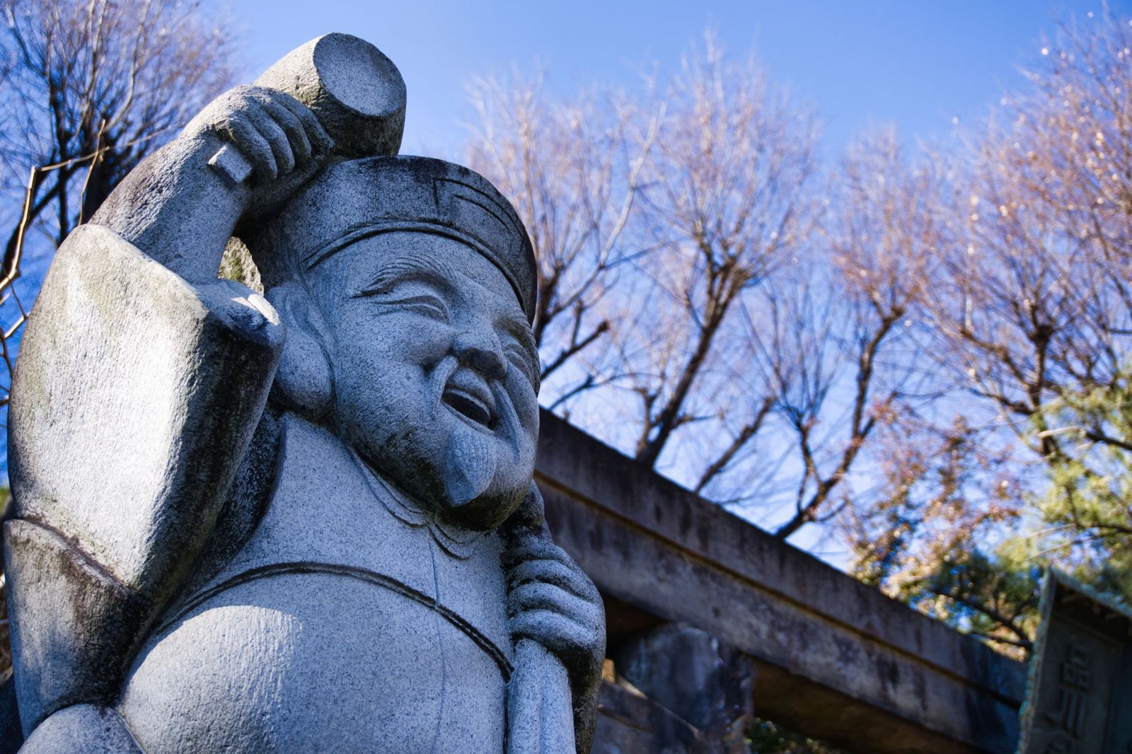 Stone statue of a joyful god holding a hammer outdoors with trees and blue sky behind it