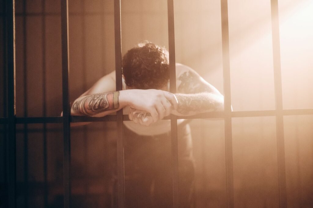 Tattooed man sitting behind prison bars with his head resting on folded arms, showing confinement and punishment