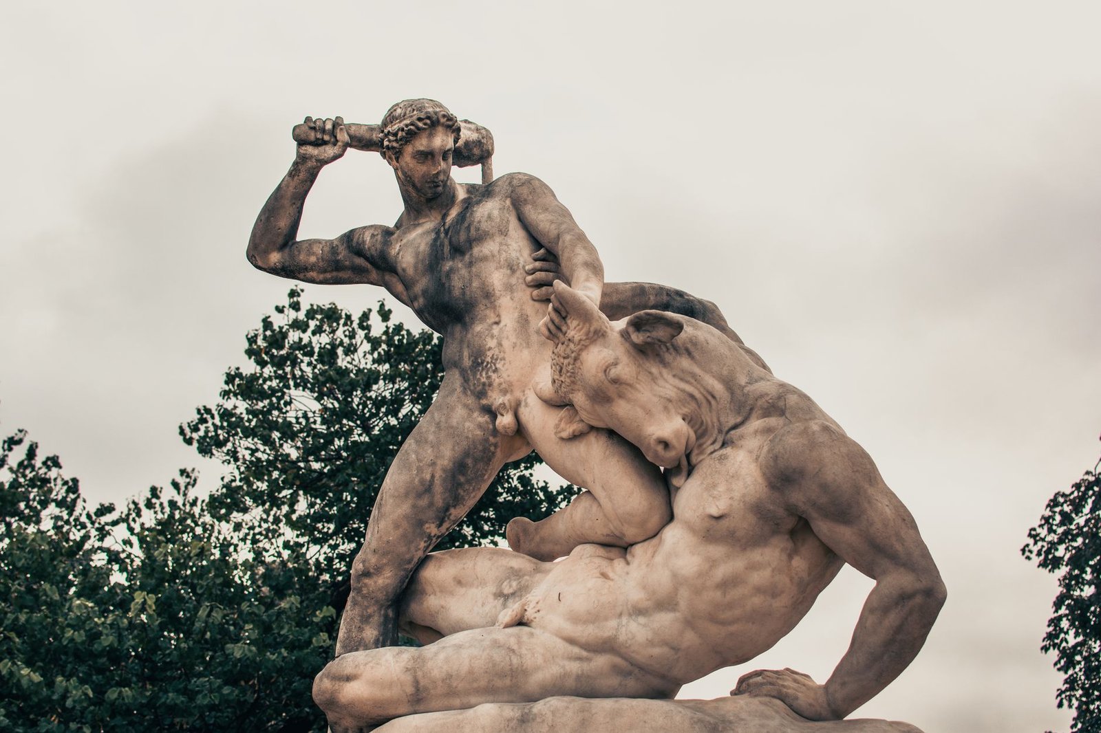 Two muscular men wrestling in a dramatic combat scene against a cloudy sky backdrop, evoking the Judgment of Paris myth