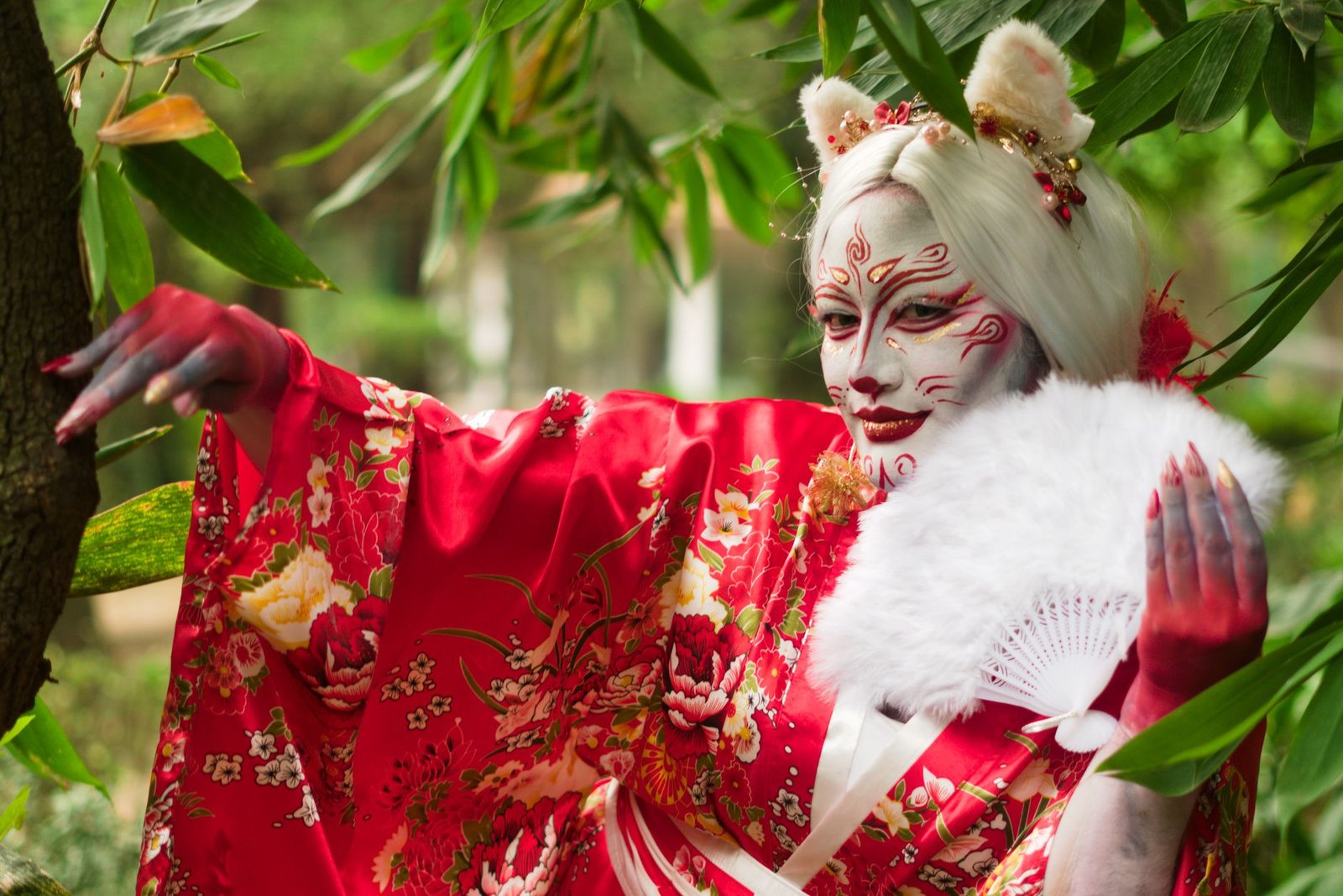 Person in red kimono with fox-inspired makeup holding a white fan in a lush green forest, evoking a kitsune spirit