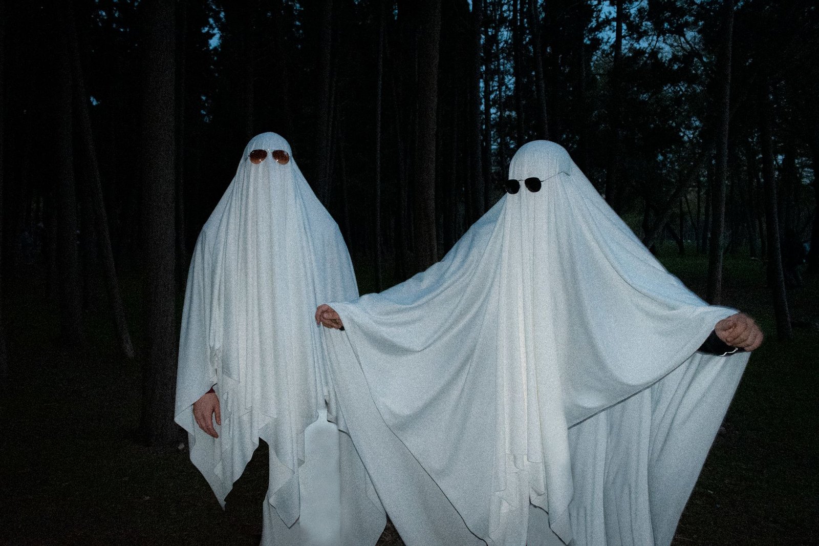 Two people in white sheet costumes stand as ghost figures in a dark forest at night