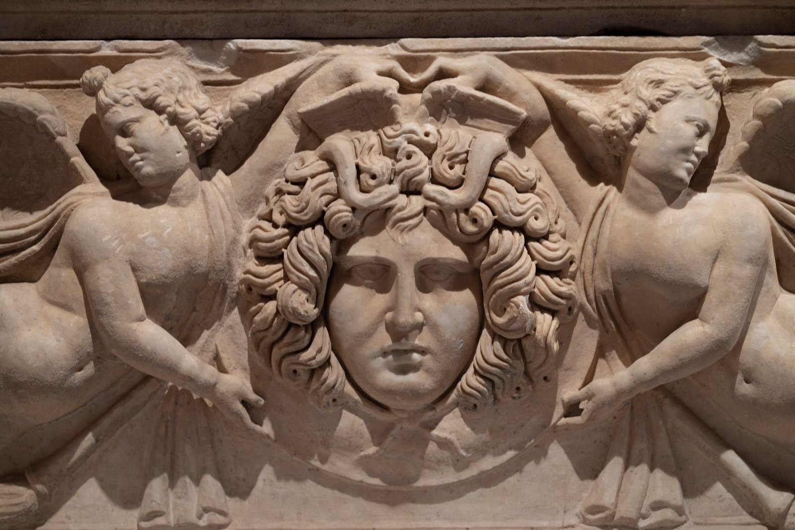 Ancient stone relief of Medusa’s head flanked by two winged figures, carved in Greek style