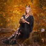 Young woman in a black witch costume sitting on a tree stump in an autumn forest, evoking the Morrigan mythological figure