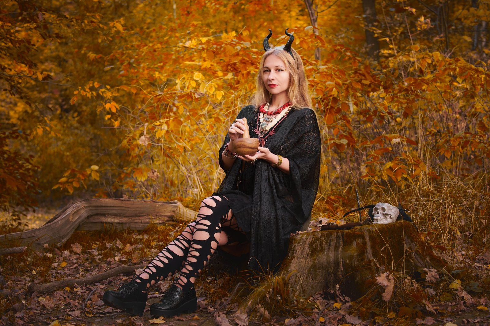 Young woman in a black witch costume sitting on a tree stump in an autumn forest, evoking the Morrigan mythological figure