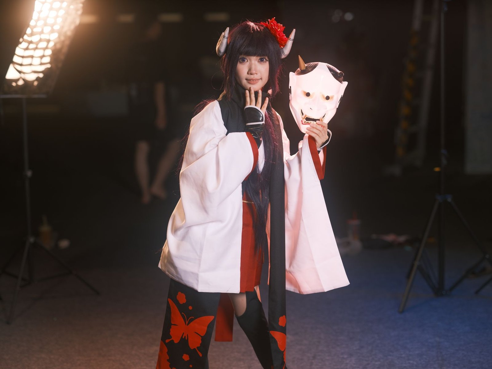 Cosplayer in traditional Japanese attire holding a white fox mask in a studio setting, evoking a tengu-inspired look