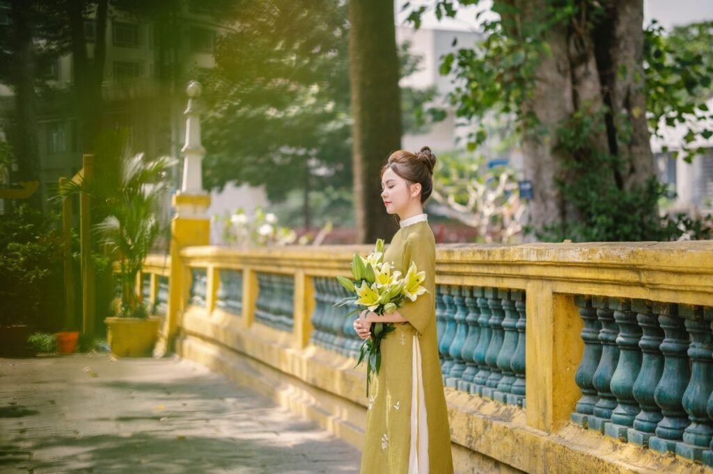 Young woman in traditional dress holding flowers beside a yellow stone railing in a serene outdoor setting, Pygmalion and Gal