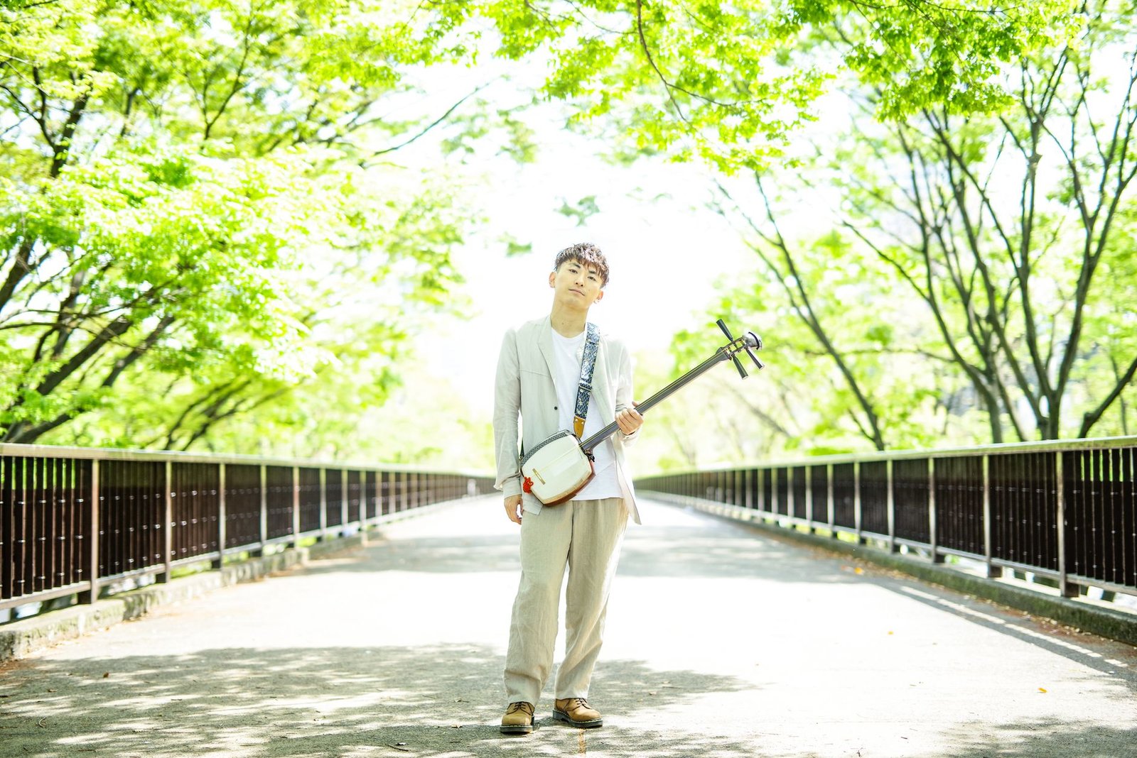 Young man playing shamisen on a sunlit bridge surrounded by green trees, depicting Sarutahiko as a guide figure
