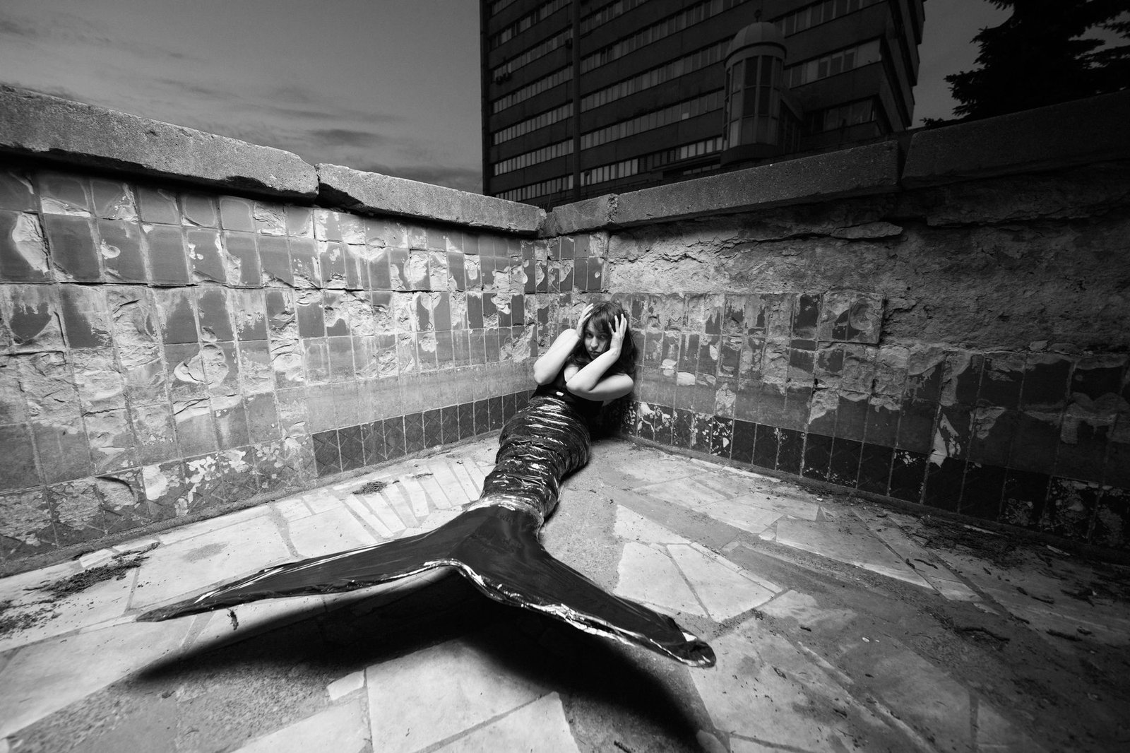 Black and white photo of a person dressed as a mermaid sitting on a tiled corner in an urban setting