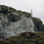 Cliffside lighthouse overlooking the ocean under cloudy skies, a modern landmark beside the legendary Lia Fáil Stone theme