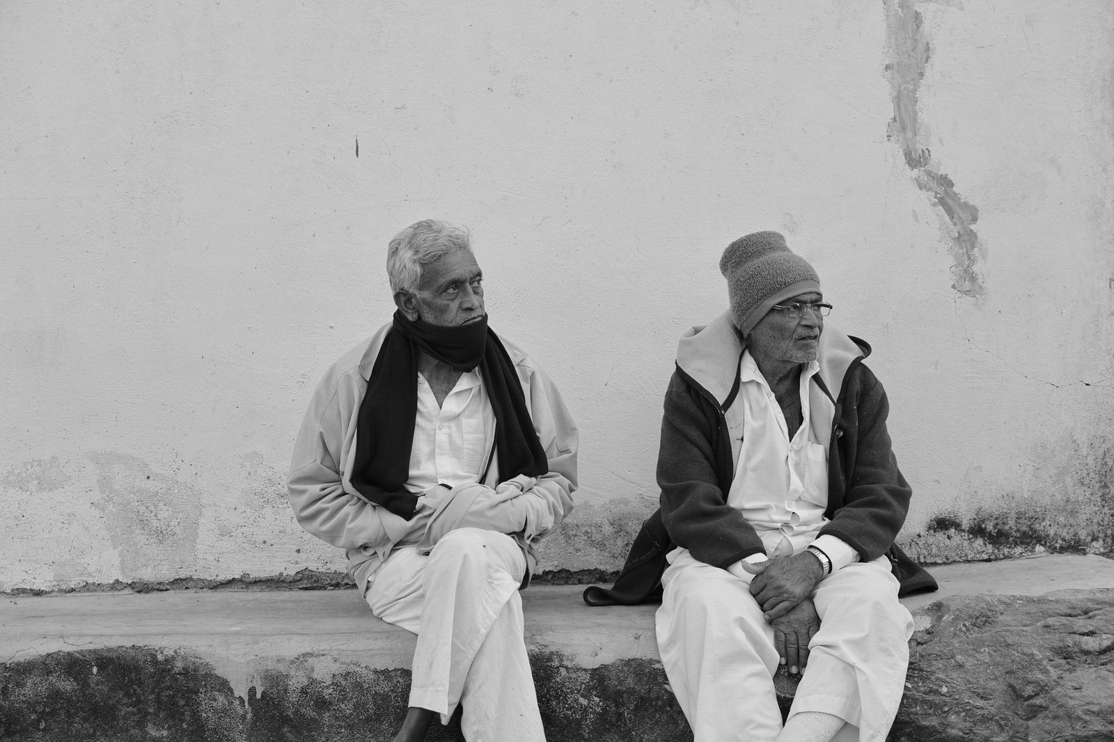 Two elderly men sit and talk on a bench against a weathered wall in black and white.