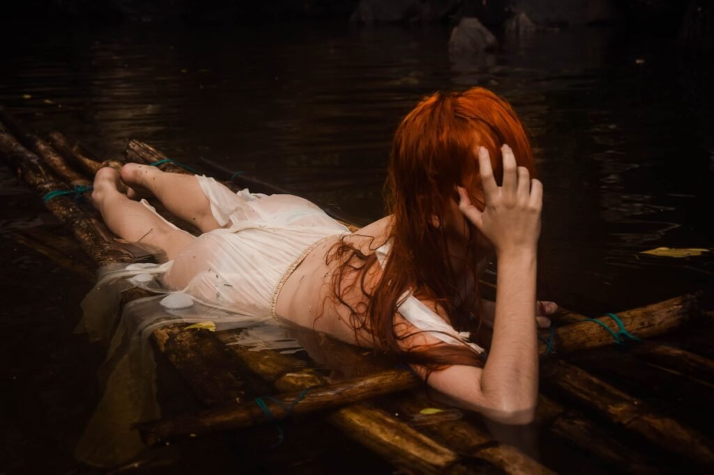 Red-haired woman lies on a wooden raft in dark water, evoking the Danaides punishment in Greek mythology