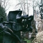 Abstract bronze sculpture beside a classical stone statue in an outdoor garden setting