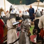 Medieval reenactors in armor and helmets clash in a battle at a historical festival, evoking the First Battle of Mag Tuired