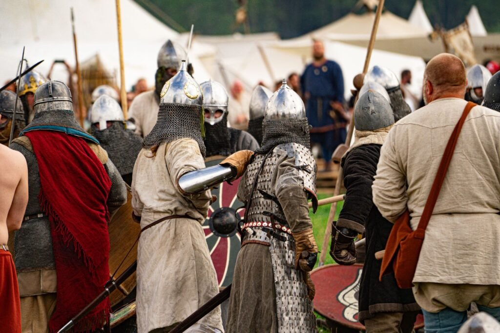 Medieval reenactors in armor and helmets clash with spears and shields at a historical festival, evoking the First Battle of