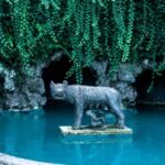 Statue of a wolf standing on a platform in a blue pond, surrounded by greenery, symbolizing Romulus and Remus