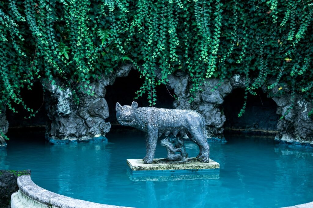 Statue of a lynx on a pedestal in a blue pond, surrounded by greenery, representing the Romulus and Remus legend