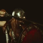Medieval warrior woman in armor holding a sword and shield against a dark background, evoking the Second Battle of Mag Tuired