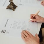 Person signing a legal contract on a white desk with a pen, representing the Roman gods Dii Consentes.
