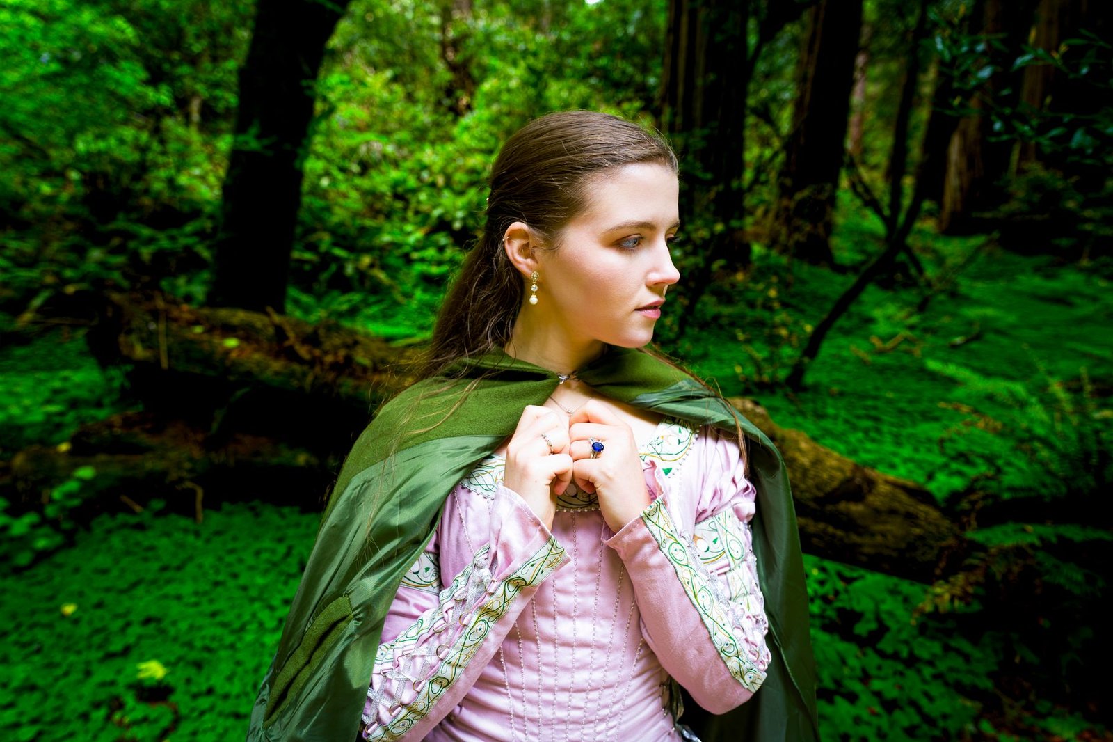 Young woman in a pink dress and green cloak standing in a green forest, representing Emer from The Wooing of Emer
