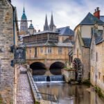 Historic European town with stone buildings and a watermill beside a river, evoking a Celtic triskele symbol legacy