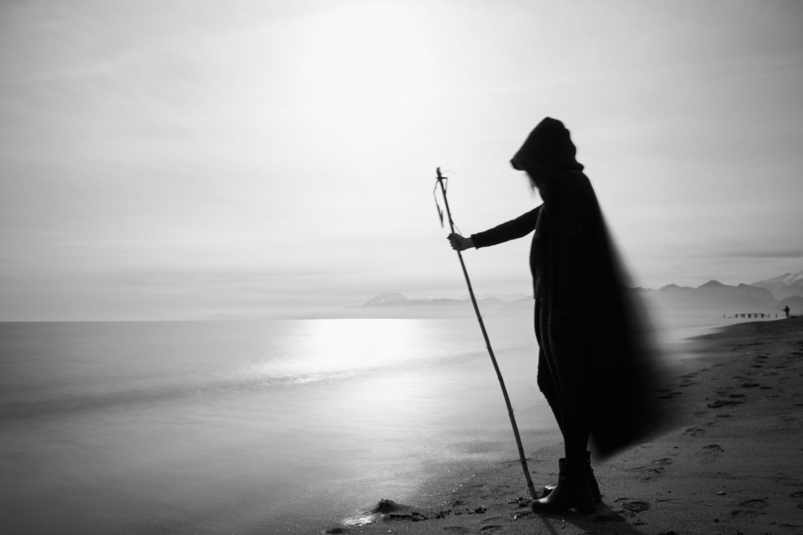 Cloaked figure holding Odin's spear on a beach at sunset, evoking Gungnir in Norse mythology
