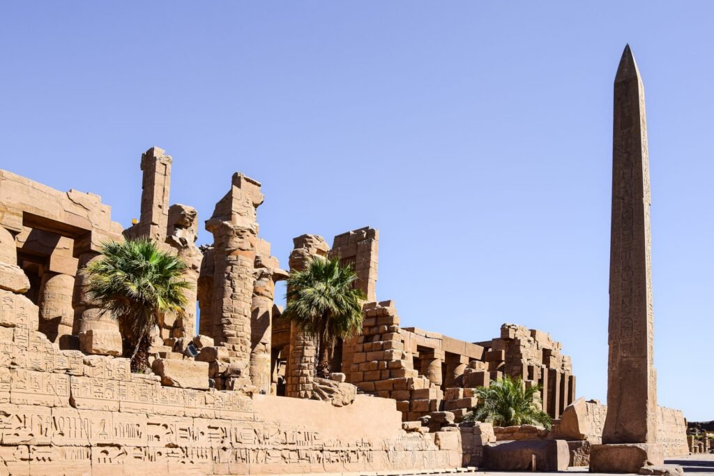 Ancient Egyptian ruins with a towering obelisk, palm trees, and clear blue sky, evoking the god Amun-Ra