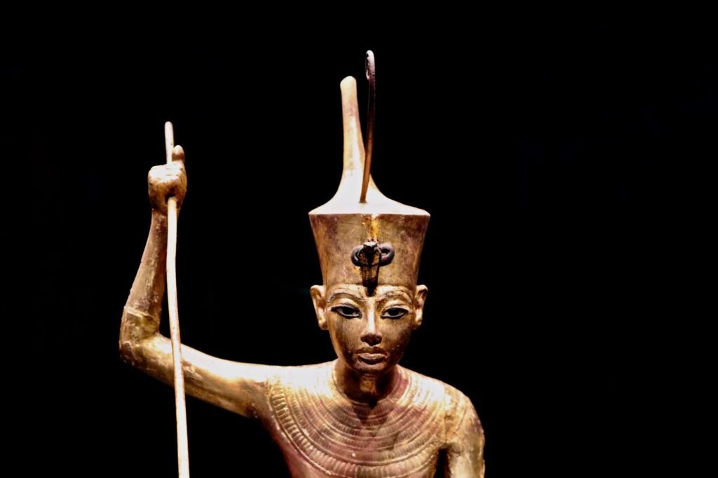 Ancient Egyptian wooden pharaoh statue holding a staff, wearing the red crown of Egypt, against a black background