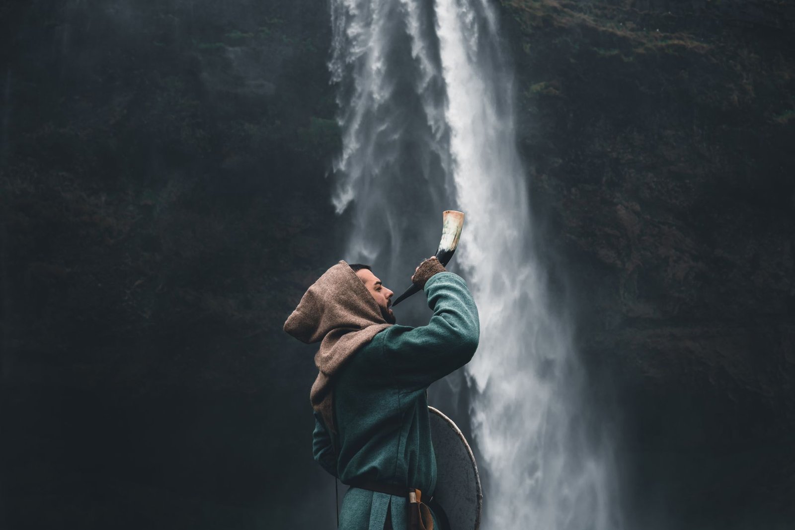 Person in a hooded jacket drinking from a waterfall, evoking the Triple Horn of Odin and the quest for wisdom.