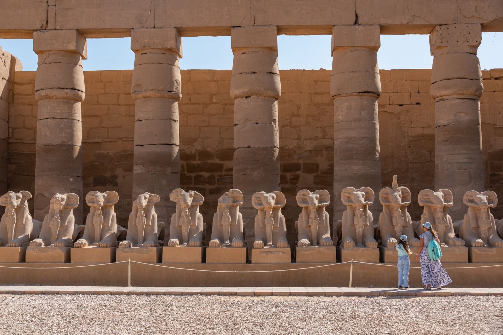 Ancient Egyptian temple with a row of ram-headed sphinx statues and tourists walking nearby