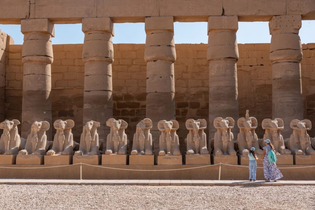 Row of ram-headed sphinx statues in front of tall stone columns at an ancient Egyptian temple site