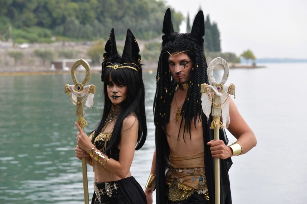 Two people in elaborate Anubis-inspired costumes holding staffs by a lakeside, with Egyptian mythology theme