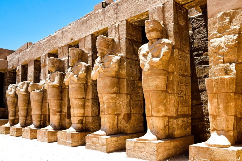 Ancient Egyptian statues of Khonsu and other figures lined up at a temple site under a clear blue sky