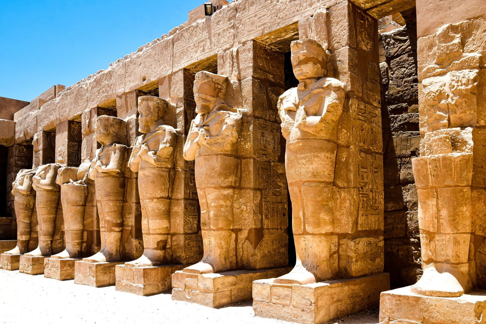 Ancient Egyptian statues of Khonsu and other figures lined up at a temple site under a clear blue sky